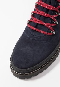 Botte de randonnée en daim bleu marine avec des lacets rouges à motif, des accessoires argentés et une semelle en caoutchouc noir texturée. Comprend des coutures blanches contrastantes.