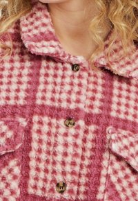 Vaaleanpunainen ja kerma-valko ruudullinen fleece-takki, jossa on kauluksellinen pääntie, suuret napit ja paikkataskut. Pehmeä tekstuuri ja lämmin ilme.