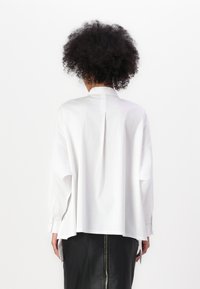 Camicia bianca a maniche lunghe con vestibilità rilassata, dotata di una piega sul retro e polsini con bottoni. Abbinata a una gonna in pelle nera con zip.
