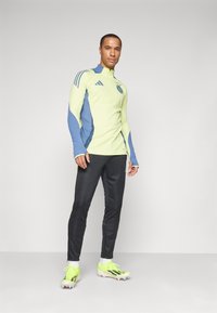 Maglietta atletica a maniche lunghe di colore verde chiaro con dettagli blu; pantaloni neri a gamba affusolata; scarpe da ginnastica neon con strisce nere; design slim fit.