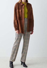 Brązowa zamszowa koszula-kurtka z dwoma kieszeniami na piersi, zestawiona z zieloną prążkowaną bluzką i czarno-beżowymi spodnie w kratę. Czarne botki na obcasie.