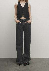 Zwart cropped vest met V-hals en knoopsluiting, gecombineerd met hoge taille, brede zwarte jeans met zakken en een klassieke denimtextuur.