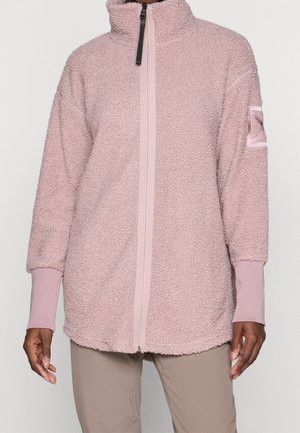 Frau trägt eine hellrosa kuschelige Fleecejacke mit Reißverschluss, hohem Kragen und langen Ärmeln, kombiniert mit beigen Hosen.