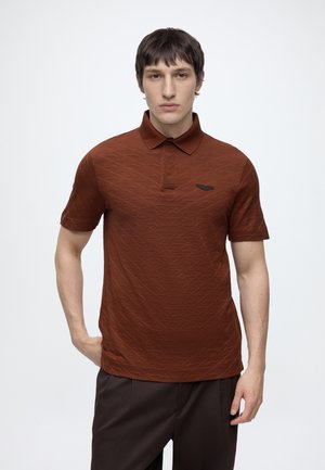 Jeune homme aux cheveux courts et foncés portant un polo marron texturé et un pantalon sombre, debout avec une main dans la poche devant un fond blanc.