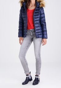 Doudoune marine avec capuche bordée de fourrure, haut rouge, jeans skinny gris, et bottes de pluie marine avec rayures et accents blancs.