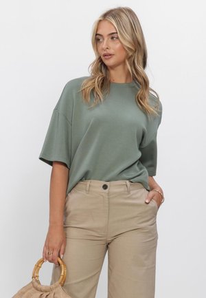 Frau mit welligem blondem Haar, die ein lockeres grünes Kurzarmhemd und beige Hosen trägt, hält eine kleine beige Handtasche mit Bambusgriffen.