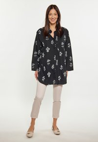 Camicia nera con bottoni, decorata con ricami floreali bianchi, maniche lunghe e vestibilità rilassata, abbinata a pantaloni cropped beige chiaro e scarpe basse neutre.