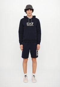 Navy hoodie and shorts set made of cotton, featuring a logo print in cream. Includes a drawstring hood and side pockets. White sneakers.