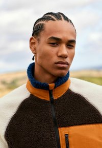 Bruine en crème kleurige fleecejas met een blauwe kraag en oranje accenten, voorzien van een ritssluiting en een voorzak.