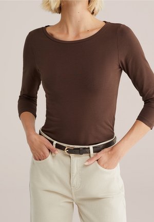 Vrouw die een aangesloten bruine lange mouw shirt draagt, ingestopt in een hoog uitgesneden crème broek met een zwarte riem, handen in de zakken, neutrale achtergrond.