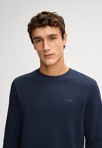 Maglione blu navy con collo rotondo e maniche lunghe. Presenta un logo discreto sul petto. Tessuto dalla consistenza liscia. Design semplice.
