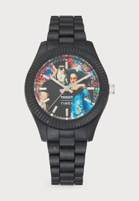 Black rubber watch with a bold patterned face featuring artwork and silver accents. Round case with embossed bezel and linked band.