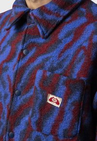 Fleece košeľa s abstraktným vzorom v červenej a modrej farbe, s gombíkovým zapínaním, špicatým golierom a náprsním vreckom s logom.