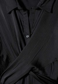 Chemise portefeuille noire avec un col, fermeture boutonnée et tissu lisse texturé. Le drapé doux crée une silhouette fluide.