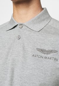 Šedé polo tričko s límečkem na dva knoflíky, měkkou texturou látky a vyšitým logem Aston Martin na levé straně hrudi.