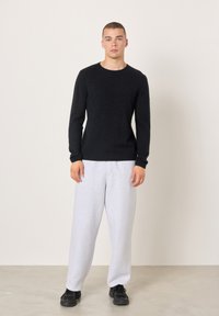 Schwarzer gestrickter Pullover mit Rundhalsausschnitt, kombiniert mit hellgrauen Fleece-Jogginghosen. Modell trägt schwarze Sneakers und steht auf einem hellgrauen Boden.