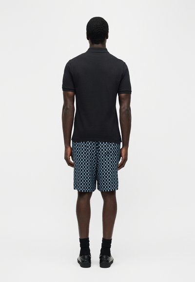 Άνδρας που στέκεται με την πλάτη στραμμένη, φορώντας μαύρο πόλο με κοντά μανίκια, μπλε σορτς με σχέδια, μαύρες κάλτσες και μαύρα παπούτσια σε λευκό φόντο.