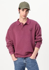 Sweat-shirt bordeaux avec un col, des poignets côtelés et un petit logo. Porté avec une casquette verte et un jeans bleu. Tissu doux et texturé.