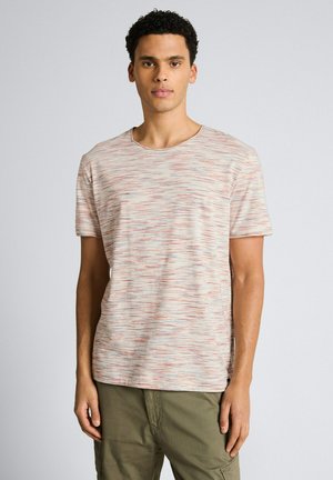 Młody mężczyzna stojący, ubrany w krótki beżowy T-shirt z cienkimi poziomymi czerwonymi i niebieskimi liniami oraz oliwkowozielone spodnie.