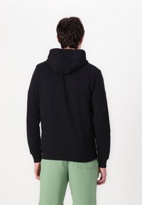 Černá mikina s kapucí se hladkým povrchem a žebrovanými manžetami, nošená přes světlé zelené tepláky, která předvádí ležérní design.