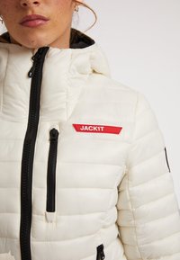 Giacca bianca imbottita con zip nera, tasche con zip e patch del logo rosso "JACK1T". Caratterizzata da un cappuccio con arricciature e texture trapuntata.