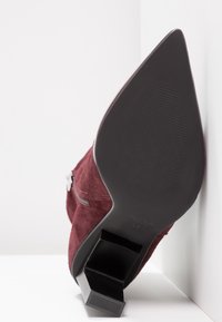 Bottine à talon haut en daim bordeaux avec un bout pointu et une semelle noire texturée, équipée d'une fermeture éclair latérale et d'un design de talon angulaire.