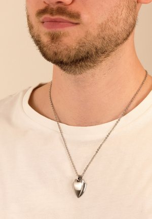 Silberne herzförmige Anhänger-Halskette an einer Kette, mit einer polierten Oberfläche und einem glatten, geschwungenen Design. Getragen über einem weißen T-Shirt.