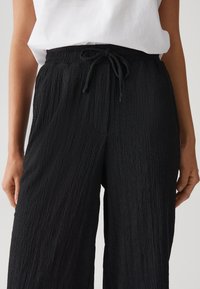 Zwarte, gekreukte stoffen broek met wijde pijpen, een elastische tailleband en een verstelbaar trekkoord. Heeft een gestructureerd oppervlak en een ontspannen pasvorm.