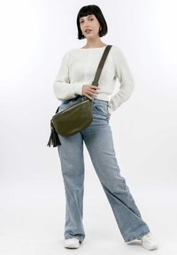 Olive green leather shoulder bag with a textured surface, zipper closure, and decorative tassels, worn with light blue flared jeans.