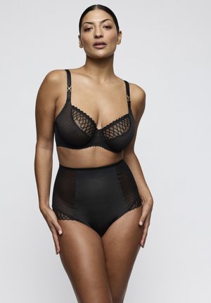 Woman modeling black lace bra and high-waisted underwear, standing against a plain light grey background with hands resting on hips.