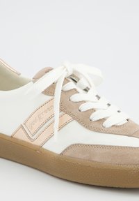 B baskets blanches avec des accents en daim beige, des lacets blancs noués en nœud, et une semelle en caoutchouc texturée marron clair.