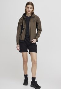 En hettejakke i brun softshellmateriale med svarte detaljer, båret over en svart skjorte og shorts. Har snorer i hetten.