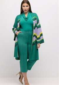 Vihreä kardigan, jossa on violetteja, keltaisia ja vihreitä raitoja, yhdistettynä yksinkertaiseen vihreään paitaan ja räätälöityihin vihreisiin housuihin. Mallilla on päällään beigetan satiini saappaat.