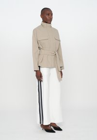 Beige belted jacket with high collar and two front pockets, paired with white wide-leg pants featuring black side stripes and black heels.