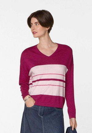 Vrouw met kort bruin haar, gekleed in een magenta en roze gestreepte V-hals trui en een donker denim rok, die een marineblauw jasje vasthoudt.