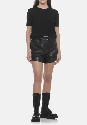 Donna che indossa una maglietta nera a maniche corte, pantaloncini in pelle neri con cintura e stivali neri con lacci fino al ginocchio, in piedi su sfondo bianco.