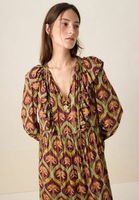 Robe longue et fluide de couleur bordeaux avec un motif floral en or et vert. Comprend un col en V, des épaules à volants et une ceinture élastique.