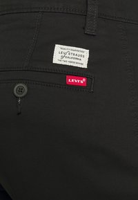 Černé bavlněné kalhoty mají přední zapínání na knoflík, malou červenou nášivku s logem Levi's a bílou textilní etiketu s detaily o značce našitou na zadní straně.