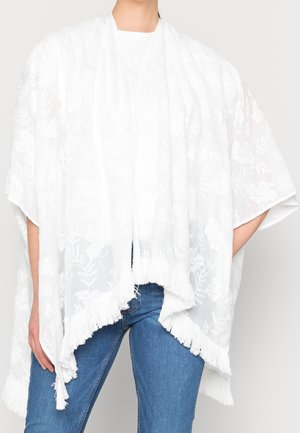 Person iført et hvidt blomsterkniplingsjal med frynset kant over en hvid top og blå jeans mod en ensfarvet baggrund.