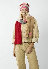 Beanie en tricot avec des rayures rose, rouge et grise ; écharpe épaisse à côtes assortie en rouge et beige. Le mannequin porte une veste utilitaire beige et un pantalon.