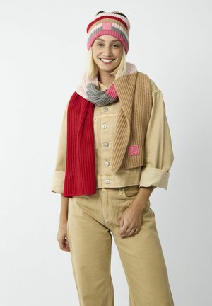 Beanie en tricot avec des rayures rose, rouge et grise ; écharpe épaisse à côtes assortie en rouge et beige. Le mannequin porte une veste utilitaire beige et un pantalon.