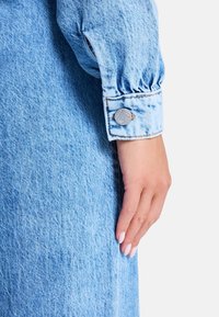 Camisa de mezclilla de color azul claro con mangas abullonadas y puños abotonados. Presenta una textura sutil y detalles de costura. El botón muestra el logo de la marca.