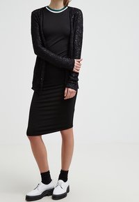 Svart cardigan med paljetter över en svart, kroppsnära klänning. Har en rund halsringning med ett kontrasterande grönt och vitt rand. Vita skor.