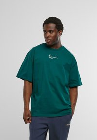 Tricou oversized teal din bumbac cu mâneci scurte, având un logo alb cu semnătura pe piept. Textură netedă și croială lejeră.