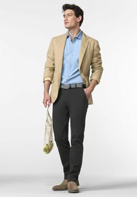 Mann in beigem Blazer, hellblauem Hemd, dunkler Hose und braunen Schuhen, der eine Netz-Tasche mit Zitronen hält und mit der Hand in der Tasche steht.