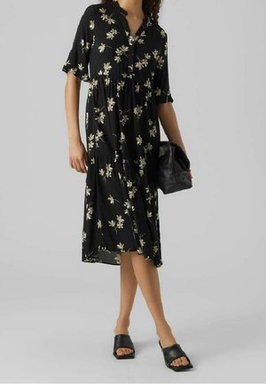 Mujer que lleva un vestido midi negro con un estampado floral claro, sosteniendo un bolso de mano negro y calzando sandalias negras abiertas en los dedos.