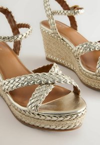 Sandales compensées métalliques dorées tressées avec brides à la cheville et talons plateforme en jute tissée sur fond clair.