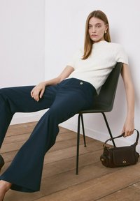 Femeie așezată pe un scaun negru, purtând un pulover alb cu mâneci scurte și pantaloni largi bleumarin, ținând în mână o geantă mică maro închis de mâner.