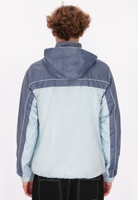 Lichtblauwe en marineblauwe windbreakerjas met een capuchon, voorzien van elastische manchetten en contrasterende stiksels langs de naden.