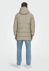 Beige Puffer-Jacke mit horizontalen Nähten, verstellbare Kapuze und mittellanger Schnitt, kombiniert mit hellblauen Jeans und weißen Sneakers.